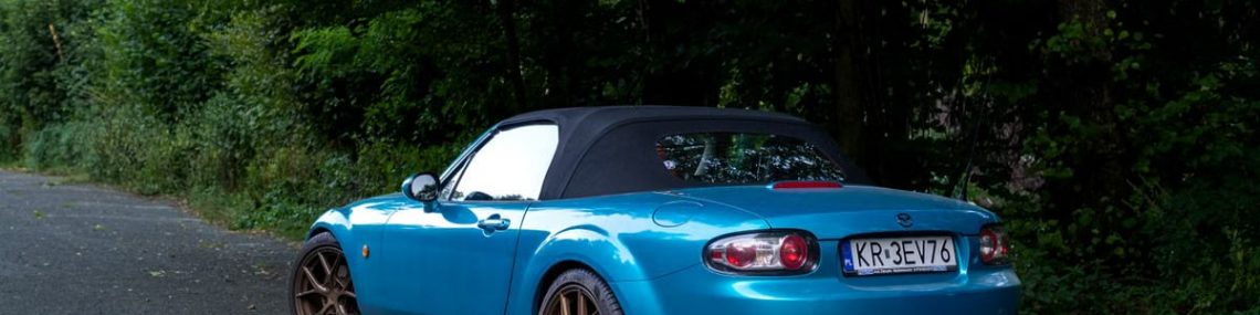 Blue Mazda MX-5 convertible parked on a forest road, showcasing its sporty design.