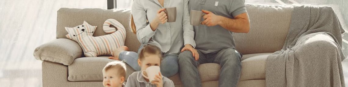Happy family with children spending time at home while parents sitting on couch with cups of coffee and looking at each other when kids resting on floor